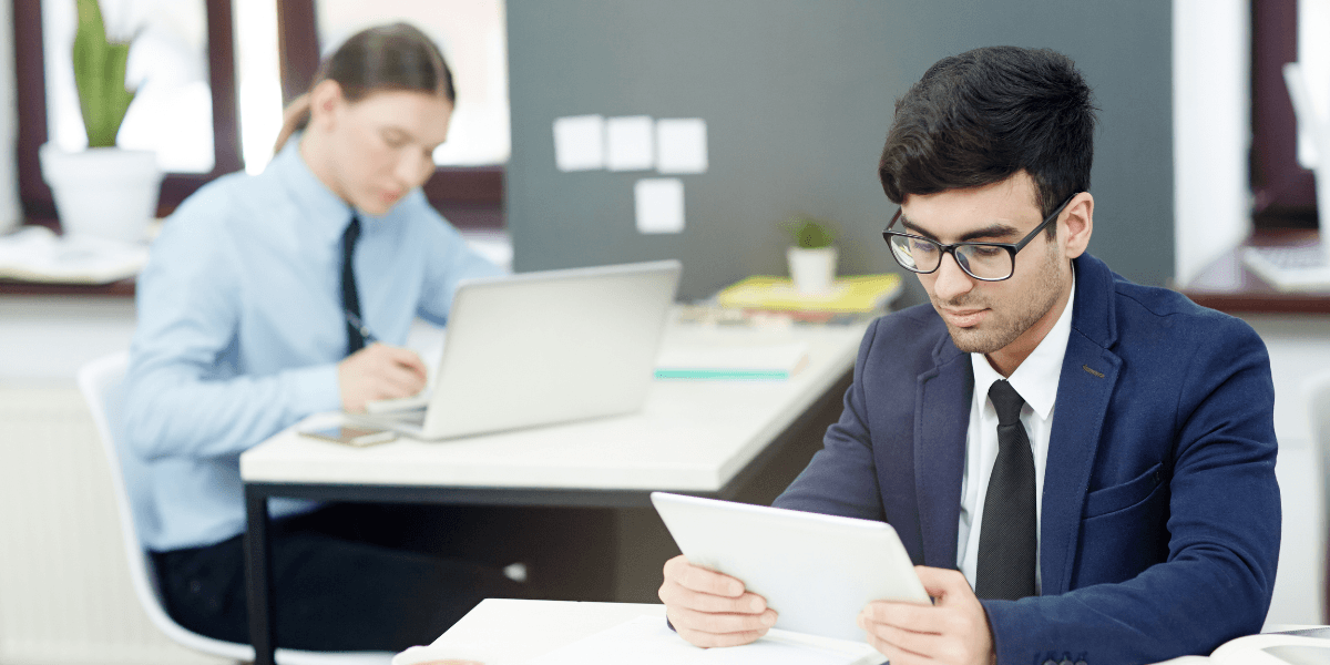 Two people work at desks in an office; one focuses on a tablet in the foreground, while the other types on a laptop in the background.
