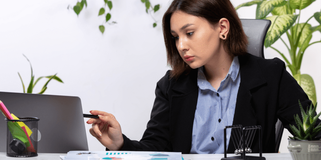 A focused woman in a black blazer works at a desk with a laptop, documents, and pen. Green plants surround the office, creating a fresh, professional atmosphere.