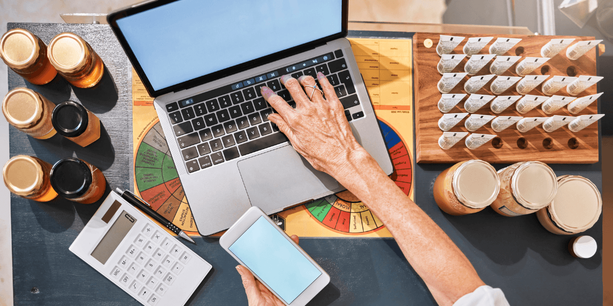 A person works on a laptop, holding a smartphone, with jars of honey and a calculator nearby. A wooden rack holds test tubes, suggesting a home business setup.
