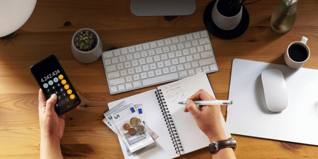 A person calculates expenses at a wooden desk, holding a smartphone with a calculator app. Nearby are Euro bills, coins, a notepad, keyboard, and coffee cup.