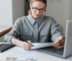 A woman in a gray shirt and glasses sits at a desk, focused on paperwork and a laptop. An American flag is placed on the desk, conveying a professional and serious tone.