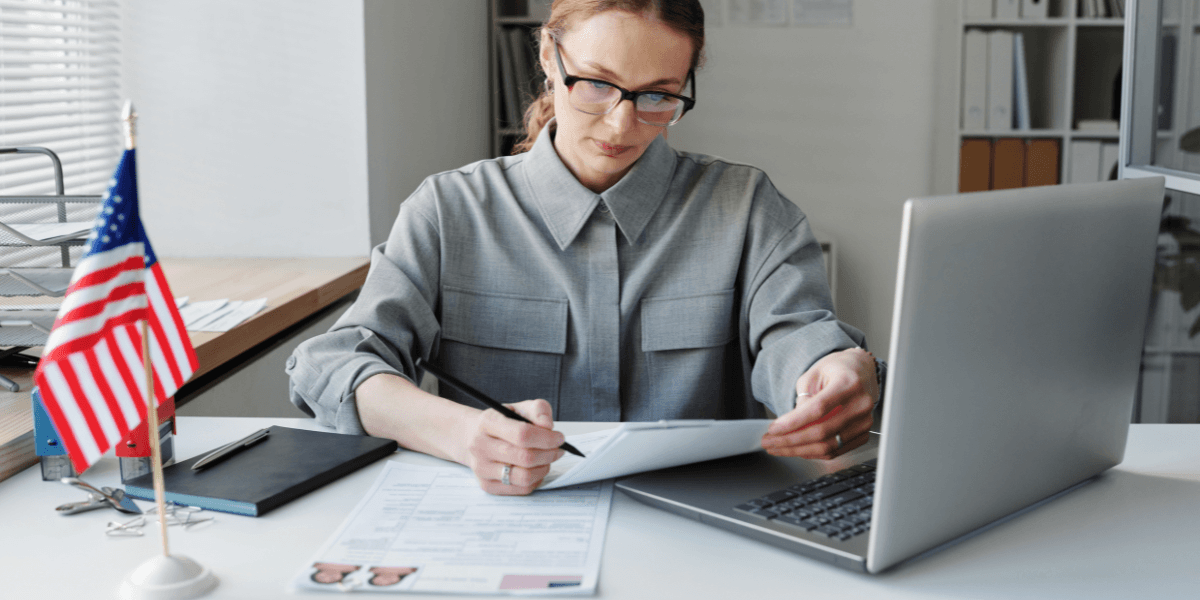 A woman in a gray shirt and glasses sits at a desk, focused on paperwork and a laptop. An American flag is placed on the desk, conveying a professional and serious tone.