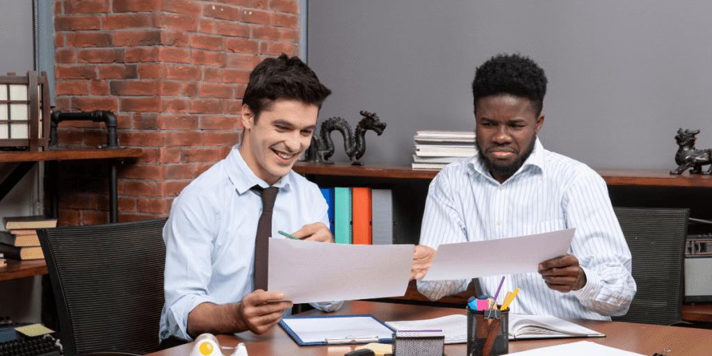 Two men in business attire sit at a desk reviewing documents; one smiles while the other looks dissatisfied. Shelves with books and figurines are visible in the background.