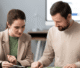 A man and woman collaborate over a laptop and papers, discussing ideas in a professional setting.