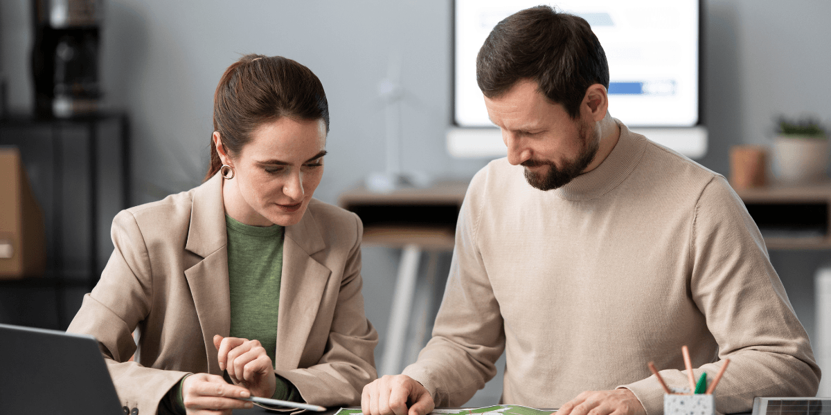 A man and woman collaborate over a laptop and papers, discussing ideas in a professional setting.