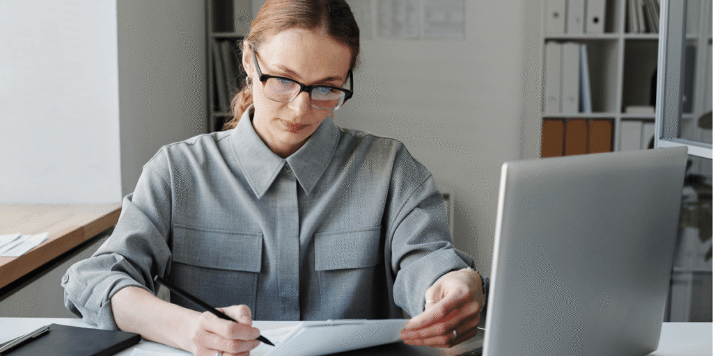 A woman wearing glasses is focused on organizing and reviewing paperwork at her desk.