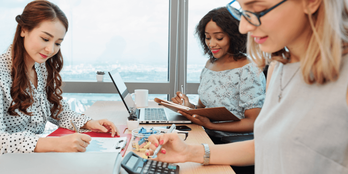 Three women collaborate at a desk, focused on a laptop, sharing ideas and discussing their project.